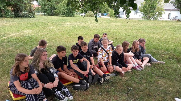 Grupa uczniów siedzi na matach w cieniu drzewa i uważnie uczestniczy w spotkaniu na świeżym powietrzu.