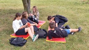 Grupa starszych uczniów odpoczywa i rozmawia, siedząc na materacach rozłożonych pod drzewem.